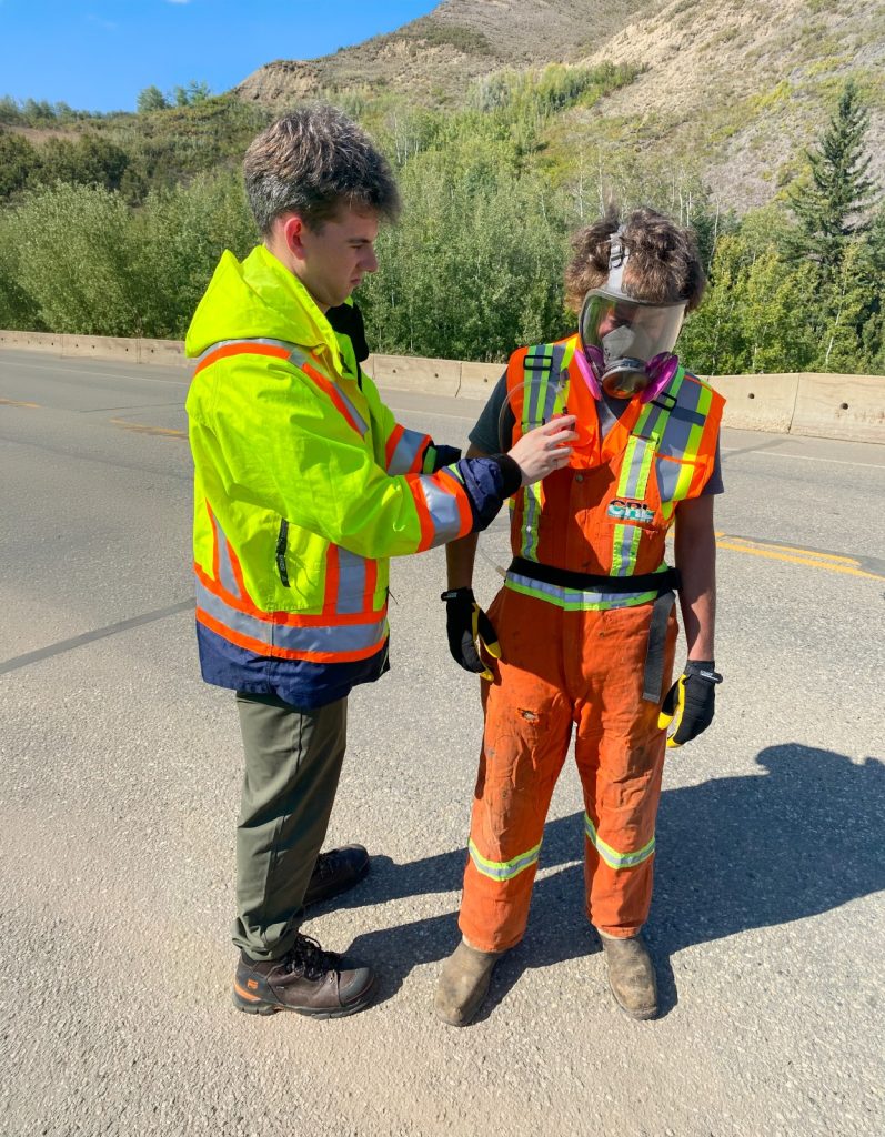 A person in a hi-vis jacket attaches an air sampling tube to the vest of a worker wearing a full-face respirator on a bridge deck.